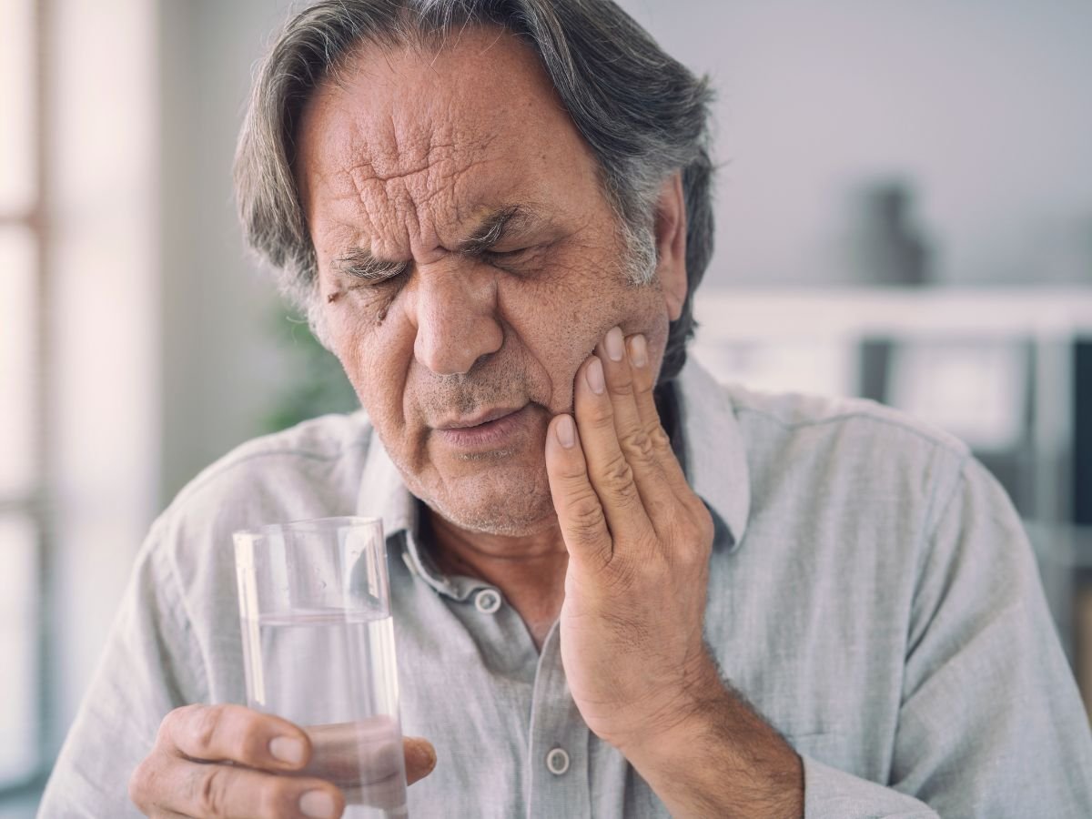 Elderly man holding a glass of water while experiencing tooth pain, showing discomfort from a possible dental issue or toothache. Oracore Dental, Urgent Dental CAre
